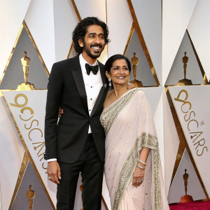 Dev Patel and Anita Patel at the Oscars event. Dev in a tuxedo, Anita in a stunning saree. The backdrop features Oscar logos and gold decorations.