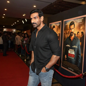 John Abraham, Indian actor, at premiere of Batla House. Wearing casual black shirt and jeans. Movie posters of Batla House visible in background. Crowded indoor event