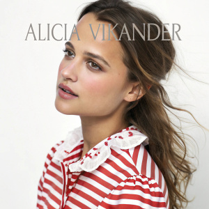 Close-up of Alicia Vikander. Wearing a red and white striped blouse with a ruffled collar. Subtle makeup and wavy brown hair. White background with text.