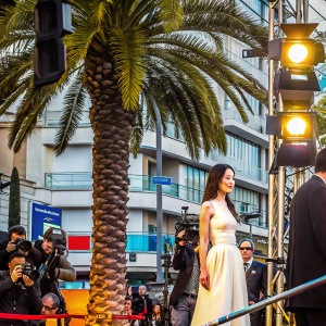 Shu Qi in a candid moment at the Cannes Film Festival. Wearing an elegant white dress. Surrounded by photographers in the bustling cityscape. Palm tree in the background