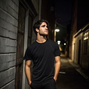 Actor Darren Criss, dressed casually in a black t-shirt, standing in front of a dimly lit alley. Solemn expression