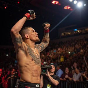 Diego Sanchez, shirtless with prominent tattoo, flexing in spotlight at UFC event. Crowd visible in background.