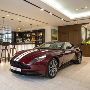 High-definition image: Classic maroon Aston Martin DB11 parked in a luxurious showroom. Elegant white stripe runs along the car's sleek body. Bright, modern interior with bar area and stylish decor.