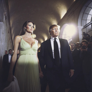 Donald Trump and Melania Trump at a glamorous charity gala. He is in a black suit with a white shirt and black tie. She is in a long, flowing, emerald green dress with a plunging neckline. Crowded venue, flashing cameras