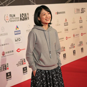 East Asian woman, friendly smile, medium-length bob hairstyle. Wearing gray hoodie and patterned black skirt. Standing on red carpet with sponsor logos. Short Shorts Film Festival & ASIA 2006 award ceremony, Tokyo.