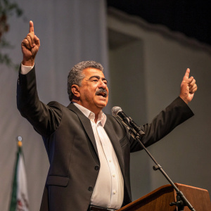 Amir Peretz, a man of Middle Eastern descent, giving an impassioned speech. Dark suit and white shirt. Stage with minimalistic backdrop and subtle decorations. Photo captures the moment of his speech, focusing on his raised hands and intense expression.