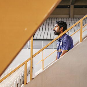 Mohammed Siraj, Indian cricketer, standing alone in stadium. Wearing blue jersey with team sponsors. Solemn expression, slight beard visible. Dim lighting, sports field in background