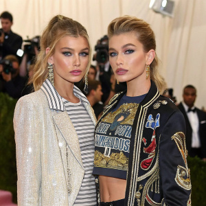Stella Maxwell and Hailey Baldwin at the MET Gala. Both dressed in avant-garde outfits, Stella in a shimmering jacket over a striped shirt, Hailey in a graphic tee with intricate designs. The backdrop is an array of vintage photographs
