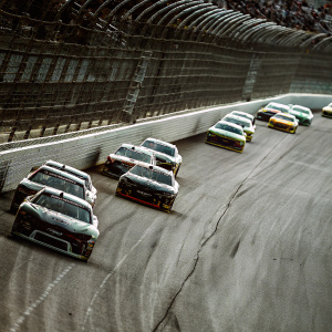 High-resolution photo showcasing a NASCAR K&N Pro Series East race. Multiple cars in fierce competition on the track. Leading car has a striking livery and visible sponsor decals.