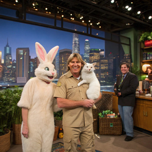 Steve Irwin in khaki outfit holding a white cat, alongside Patricia Arquette in a fluffy rabbit costume. Jay Leno and Miguel Ferrer also present. The scene takes place in a bustling studio with a cityscape backdrop.