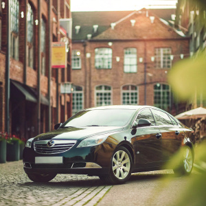 In a vibrant courtyard, a shiny Vauxhall Insignia EXCLUSIV NAV CDTI ECOFLEX stands out in sleek black. Surrounding it, an array of buildings are marked with various signs and banners. Sunlight bathes the area in a warm golden hue.