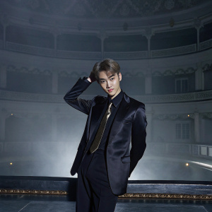 Kim Se Hun, EXO-K member, in a sleek black suit with a subtle gold tie pin, posing confidently on stage at a grand concert hall. Dim lighting highlights his fair skin and sharp features.