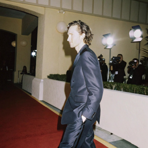 Tom Hiddleston, wearing a sleek black suit, attending the Golden Globe Awards. The venue is filled with red carpet, flashing lights and cameras.