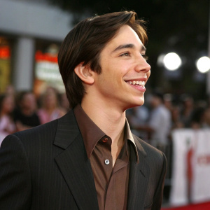Justin Long at the movie premiere of DodgeBall: A True Underdog Story in Westwood, California. Wearing a dark suit over a brown shirt, with styled hair and a cheerful expression. Blurred audience in the background.