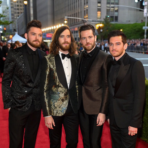 Dan Smith, Chris Wood, Kyle Simmons, and Will Farquarson of Bastille at the MET Gala. They are dressed in extravagant evening wear. A bustling New York City street in the background.