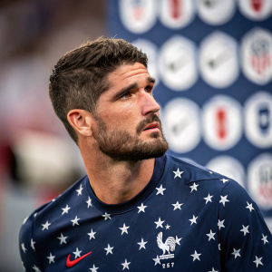 Professional soccer player Rodrigo De Paul. Concentrated face, trimmed beard, in a close-up shot. Dark blue jersey with white stars. The blurred background features numerous logos.