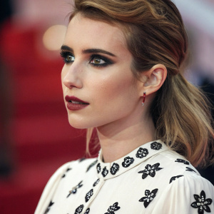 Emma Roberts in an intense close-up. Dramatic makeup with dark eyes and bold lips. She is in a white blouse with black patterns. Blurry red background, only her face and upper body visible.