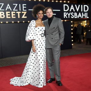 Zazie Beetz and David Rysdahl at a glamorous award ceremony. Zazie Beetz is wearing a stunning white polka dot dress with a voluminous top layer. David Rysdahl is in a sleek gray striped suit. They stand on a red carpet with illuminated logos and words behind them.