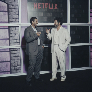 Adam Sandler and Luis Gerardo Mendez at the Netflix launch party. They stand in front of a large, vibrant backdrop filled with colorful graphics and text.
