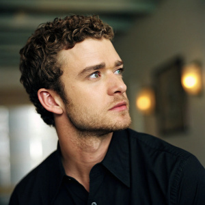 Close-up portrait of Justin Timberlake. Wearing a black shirt. Curly hair and stubble. Indoor setting, blurred background