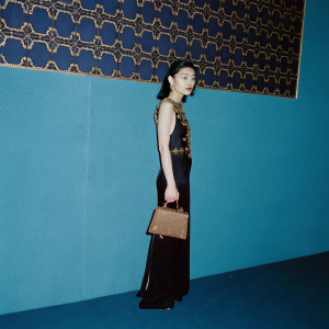 Maggie Cheung at a fashion gala. She is wearing a sleek black dress with gold embellishments and a brown structured clutch. The backdrop is a geometric patterned wall.