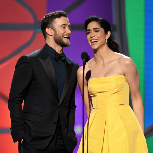 Justin Timberlake and Sarah Silverman on a vibrant stage. Justin, in a sharp black suit, grins beside Sarah, dressed in an eye-catching yellow gown, at a comedy gala.