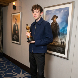 Alex Lawther, Caucasian male, standing with Oscar statuette in hand. Formal attire, dark blue jacket, black pants. Movie poster backdrop.