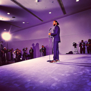 Colin Kaepernick, wearing a navy blue suit, speaking at a press conference. Dark stage with bright lights