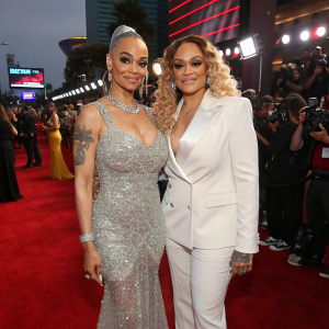 Charli Baltimore and Faith Evans at a glamorous awards show. Both wearing stunning outfits. Charli in a silver sequined gown, Faith in a white tailored blazer. Crowded and vibrant red carpet setting.