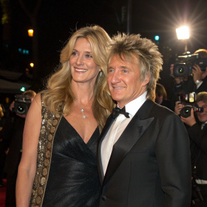 Rod Stewart and Penny Lancaster at a glamorous Hollywood party. Rod, in a sleek black tuxedo, and Penny, with her golden locks flowing, pose together under flashing cameras.