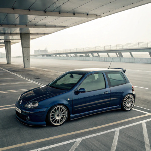 A sleek, dark blue Renault Clio V6 Sport hatchback, with sharp angles and smooth curves. Silver wheels, glossy black windows. Empty, futuristic parking lot