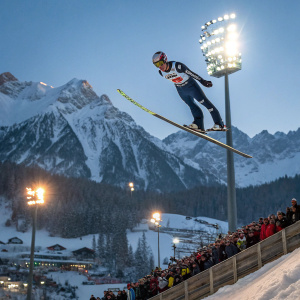 Kamil Stoch mid-air at a winter sports festival. Bright lights and snowy peaks visible. Spectators in the background