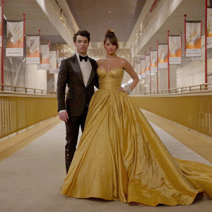 Kevin Jonas and Danielle Deleasa at an awards show. Both in stunning formal wear. Danielle in a strapless gold gown. Background: starlit sky with event banners.