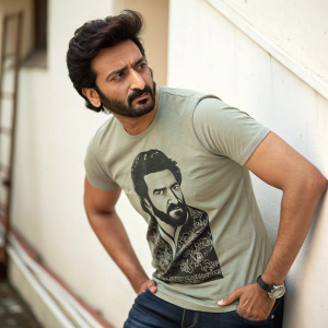 A man confidently posing with a beard and strong jawline, wearing a t-shirt featuring a detailed graphic of a Bollywood actor. Simple, plain background.
