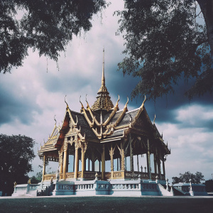 The Barom Mangalanusarani Pavilion in Ananta Samakhom Throne Hall, Bangkok, Thailand. Ornate structure with multiple levels and intricate gold detailing. Traditional Thai architecture with elaborate carvings. Surrounded by a grassy area and statues, under a partly cloudy sky.