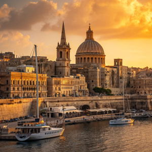 Valletta, Malta's capital, captured at sunset. Golden hues bathe the historic buildings. Boats docked along the waterfront. Reflective water mirrors the bustling cityscape. Diverse architecture, from ancient to modern, with intricate balconies.
