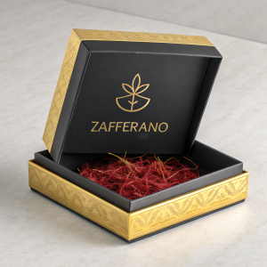 Premium saffron packaging. Sleek black and gold box. 'ZAFFERANO' logo prominently displayed. Inside view of red saffron. Neutral background