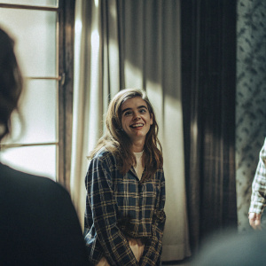 Kiernan Shipka, in a candid photo shoot, displays a playful grin with her hair styled into waves. She wears a plaid shirt. The backdrop is a blurred curtain with soft lighting.