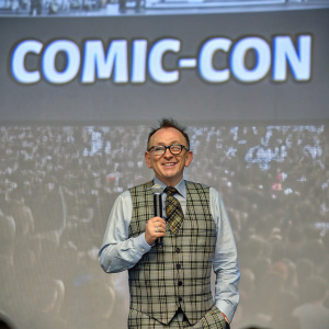 Michael Emerson, actor, standing in front of a bustling fan convention. Dressed in a plaid vest, shirt, tie, and glasses. Holding a microphone. Graphic backdrop with Comic-Con text. Energetic crowd in the background.