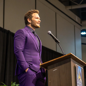 Chris Pratt at a galactic convention. Purple suit, standing at podium. Microphone in hand. Indoor stage, simple backdrop