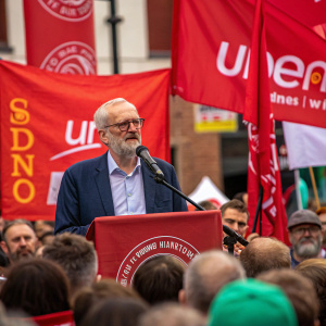 Jeremy Corbyn addressing a rally. Bearded, glasses, center stage. Audience in the foreground. Event sponsored by union. Red banners and union logos prominent.