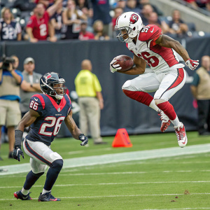 Sports moment captured: Houston Texans player in #26 crouching stance, Arizona Cardinals player in #12 airborne leap. Spectators in background.