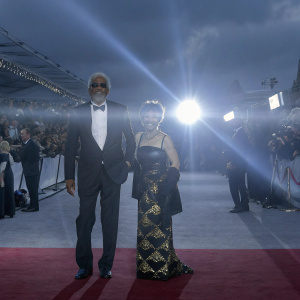 Morgan Freeman and Rita Moreno at the Oscars. Freeman in a black tuxedo, Moreno in a black gown with gold accents. Crowds, photographers, and a red carpet backdrop.