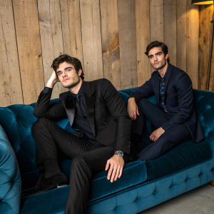Jacob Elordi and a fellow actor, both dressed in dark suits, posing on a luxurious blue velvet sofa. Rustic wooden wall backdrop with warm lighting