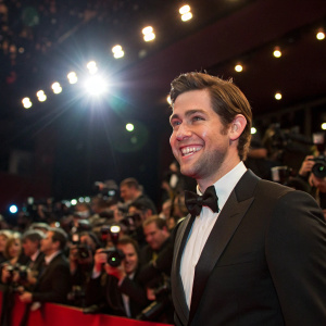 John Krasinski, wearing a black suit and bow tie, smiles at the premiere of his latest film. Crowded theater with flashing cameras