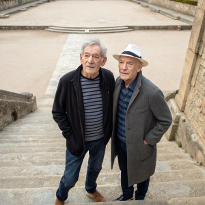 Sir Ian McKellen and Patrick Stewart, both in casual attire, standing on ancient stone steps. Patrick Stewart wears a dark jacket and striped shirt, while Ian McKellen is in a gray jacket with a white hat. The background is a quiet, empty courtyard.