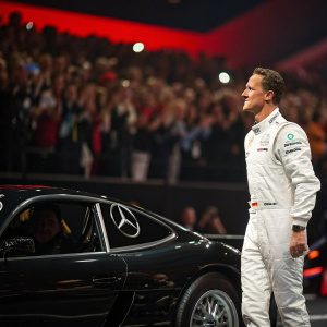 Michael Schumacher, intense focus, dressed in white racing suit. Standing beside sleek, black racing car. Event: Sports Award Ceremony. Audience cheering in the background.