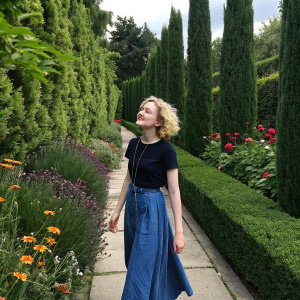 Julia Garner in casual attire, black t-shirt and blue skirt, amidst a vibrant garden with tall green hedges.