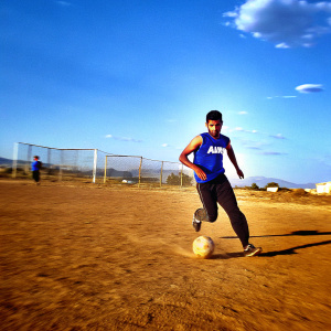 Photograph of Andreas Pereira, soccer player, intensely focused. Running on a soccer field. Wearing blue sleeveless jersey with AON logo, black pants. Background blurred, creating depth.