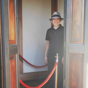 David Archuleta, young Hispanic male, smiles brightly at the camera. Wearing a black polo shirt and a fedora hat. Standing behind a red velvet rope. Warm, natural lighting